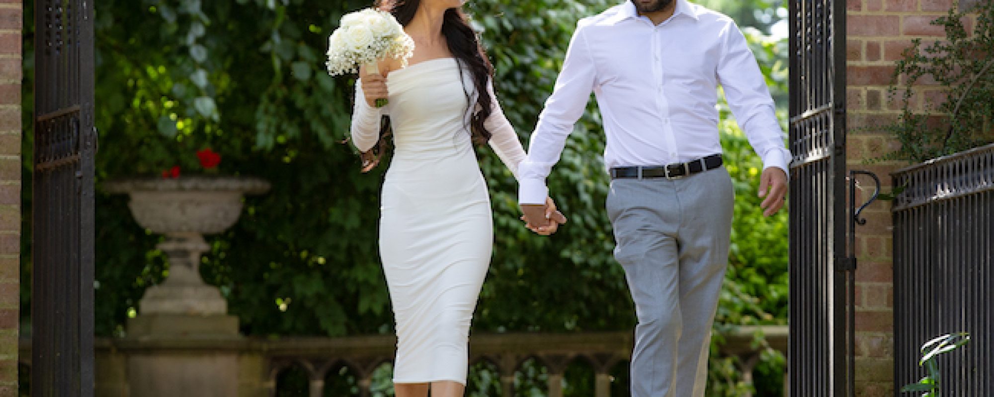 Bride and Groom walking through black metal gates into a walled garden in the sunshine looking at each other